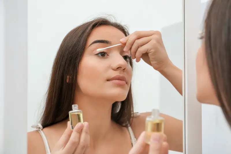 beautiful young woman removing eyelash glue by applying oil to her eyelashes near a mirror